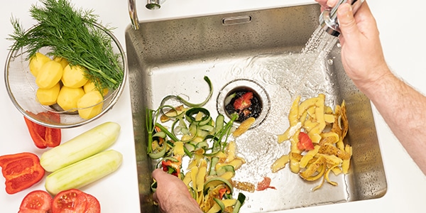 stainless steel sink with vegetables in sink and hand holding sprayer as vegetables go down garbage disposal